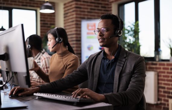 African american helpline employee working at call center reception with multiple monitors. Male operator using telecommunication to help clients at customer service support, remote network.